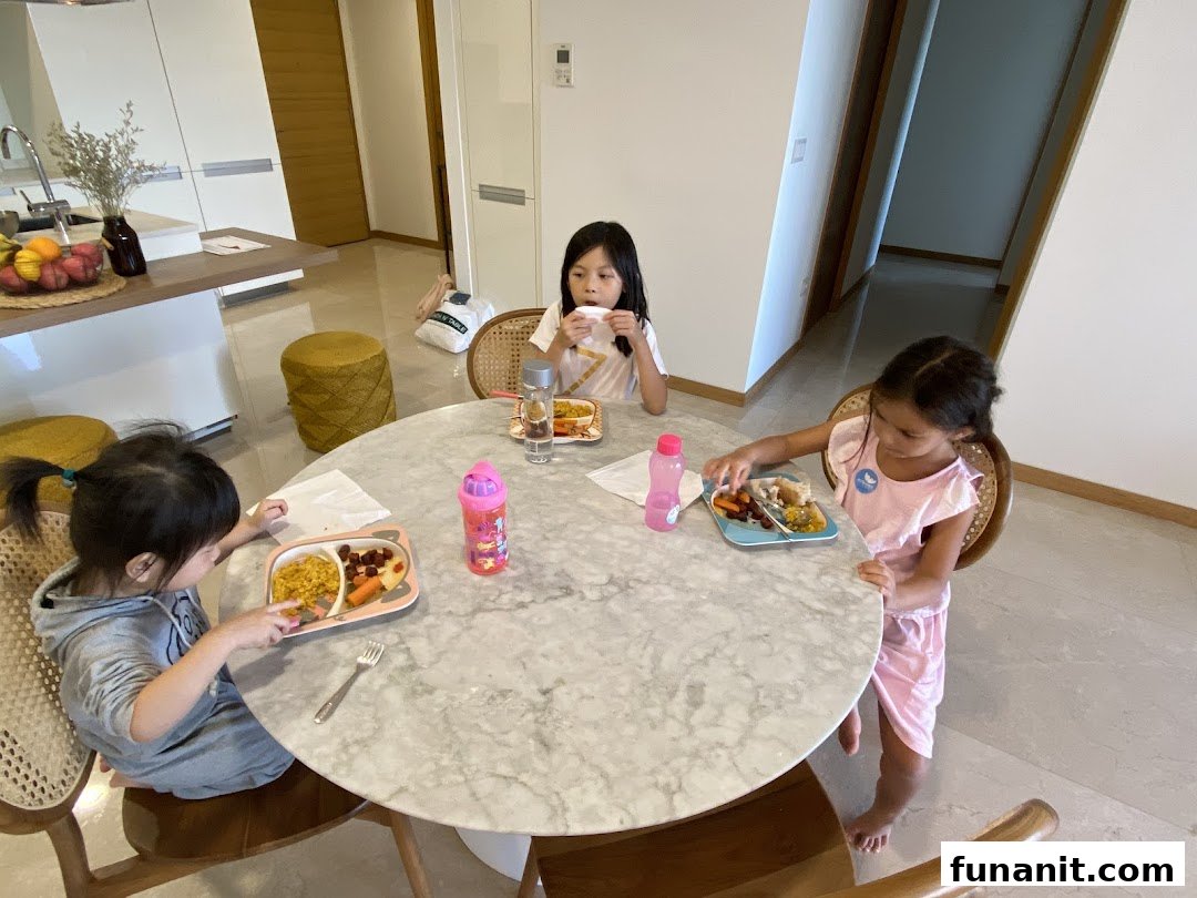 Three children are seated around a round table, eating meals in a bright home kitchen setting.