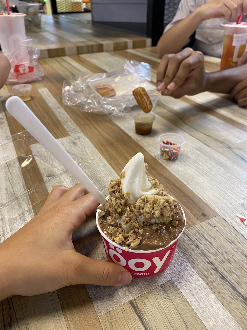 A hand holds a smöoy frozen yogurt cup with toppings, alongside churros and dips on a wooden table.