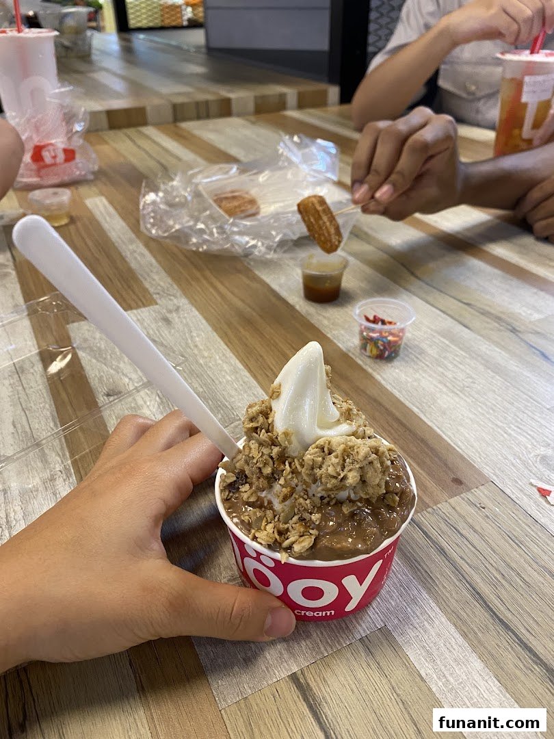 A hand holds a smöoy frozen yogurt cup with toppings, alongside churros and dips on a wooden table.