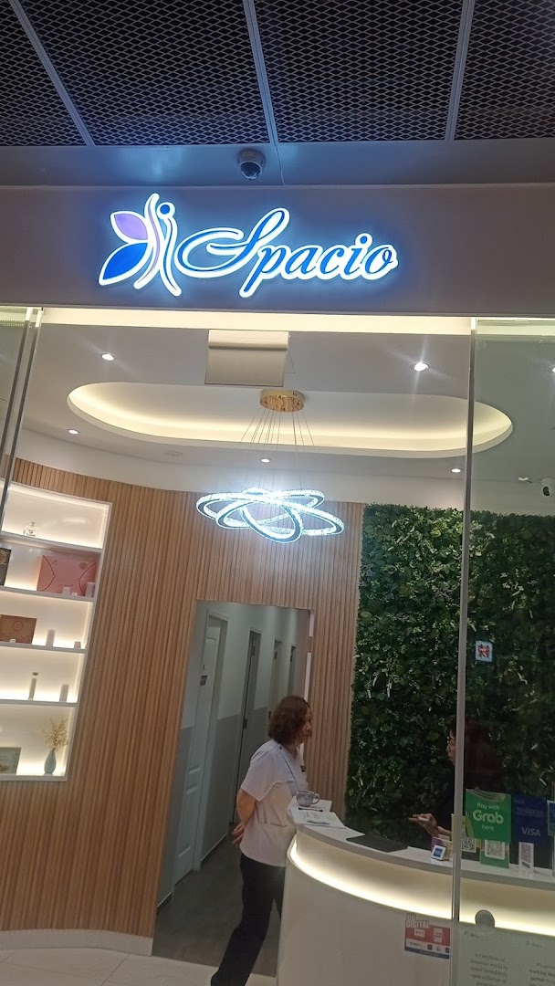 The image shows the bright, modern reception area of Spacio, featuring its illuminated logo and a green wall.