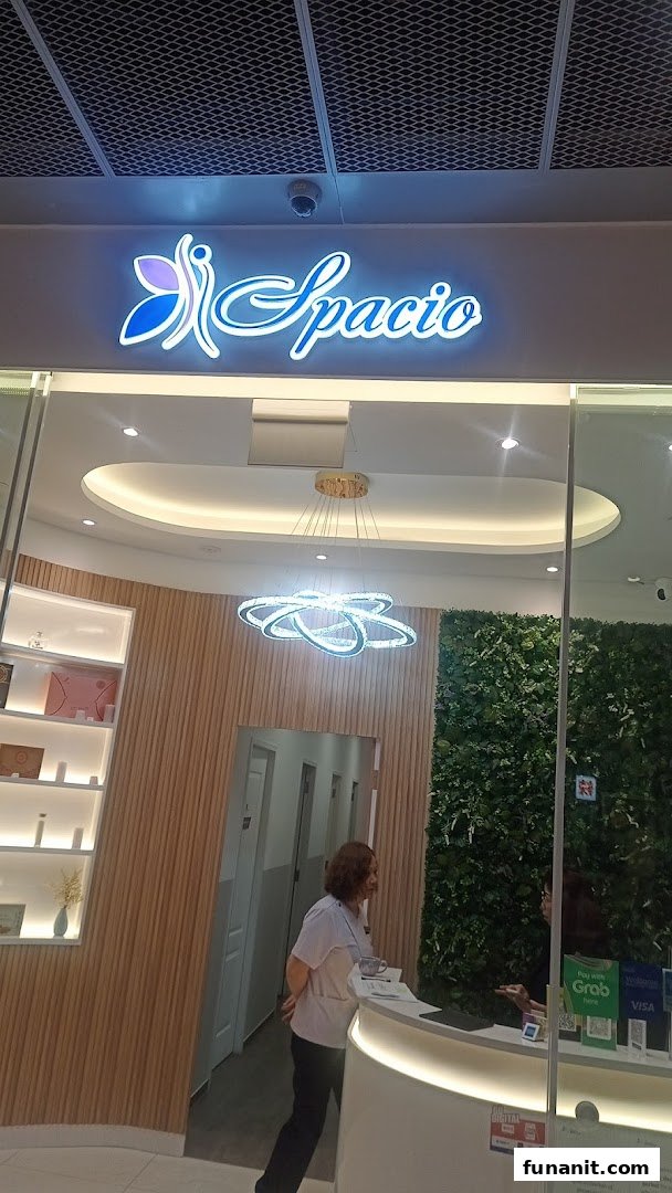 The image shows the bright, modern reception area of Spacio, featuring its illuminated logo and a green wall.