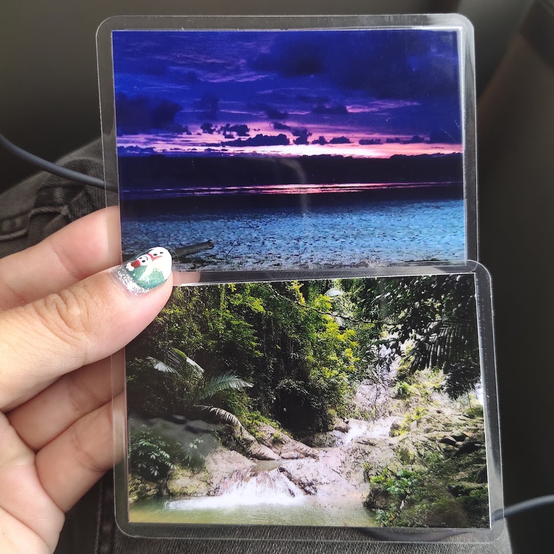 A hand holding two laminated photos: a vibrant sunset over water and a lush waterfall scene.