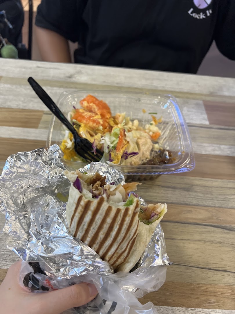 A hand holding a fresh wrap in foil, with a colorful salad bowl and fork on a wooden table.