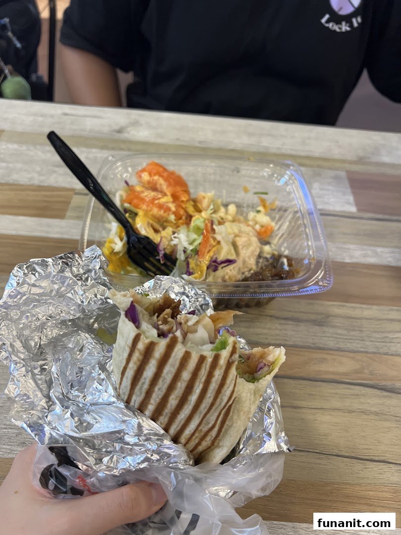 A hand holding a fresh wrap in foil, with a colorful salad bowl and fork on a wooden table.