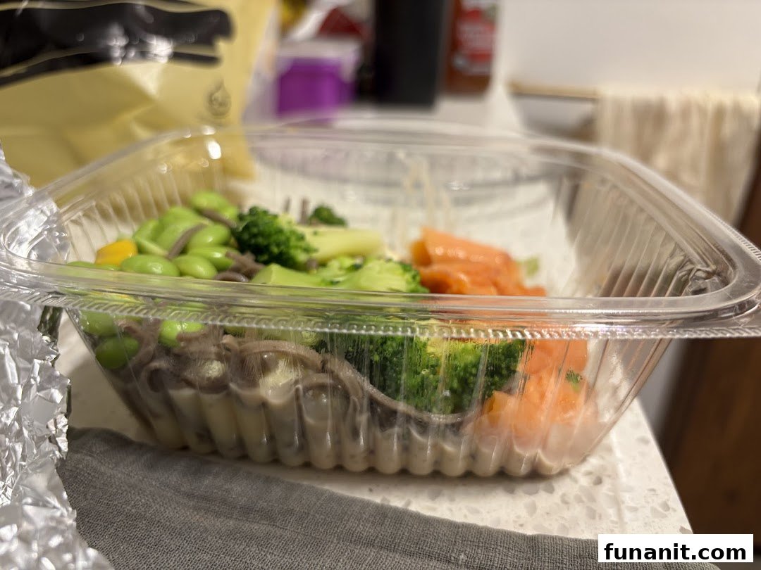 A healthy meal in a clear container featuring edamame, broccoli, noodles, and salmon.