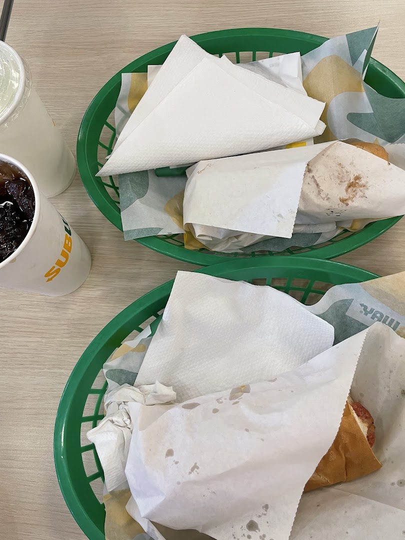 Two Subway sandwiches in green baskets with drinks on a light wooden table.