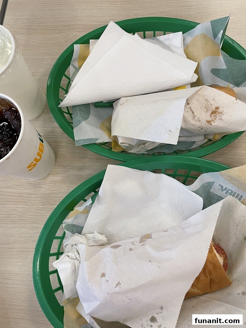 Two Subway sandwiches in green baskets with drinks on a light wooden table.