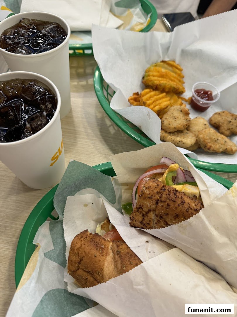 Two Subway sandwiches, waffle fries, chicken nuggets, and drinks on a table.