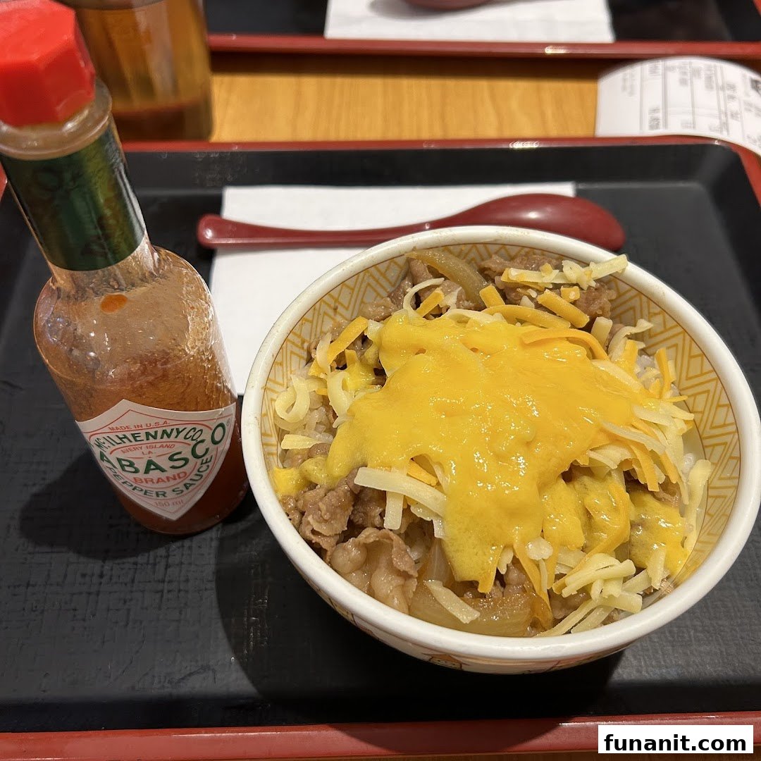 A close-up of Sukiya's gyudon bowl topped with melted and shredded cheese, served with Tabasco sauce.