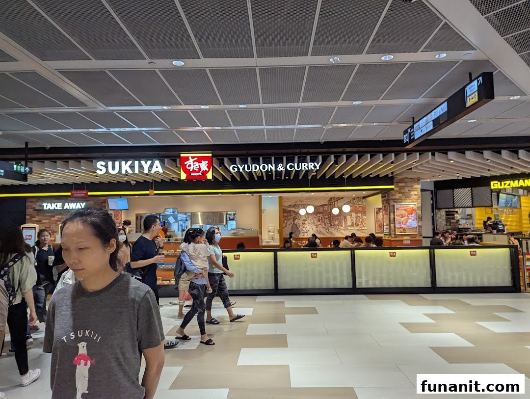 An interior view of Sukiya Gyudon & Curry in a bustling mall food court, with customers and staff visible.