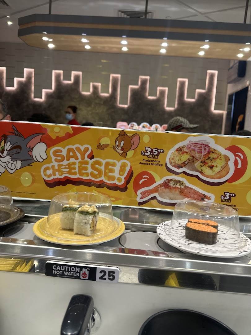 Conveyor belt sushi restaurant interior featuring plates of sushi, a Tom and Jerry banner, and a hot water dispenser.