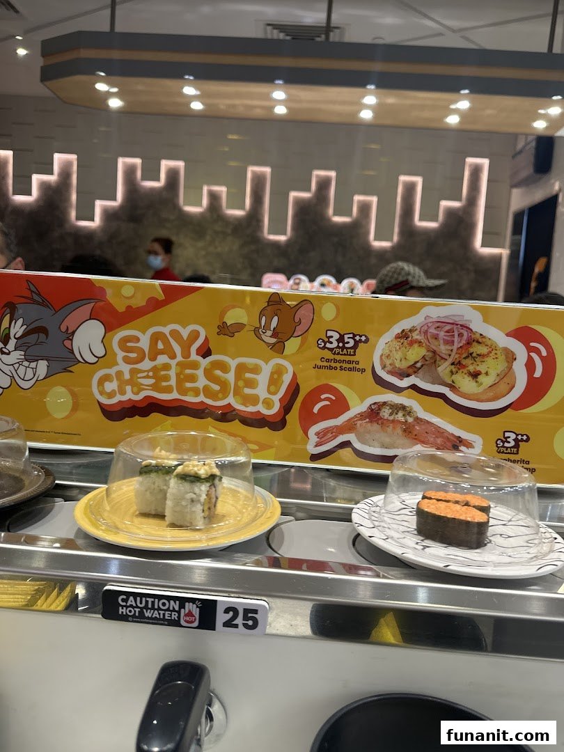 Conveyor belt sushi restaurant interior featuring plates of sushi, a Tom and Jerry banner, and a hot water dispenser.