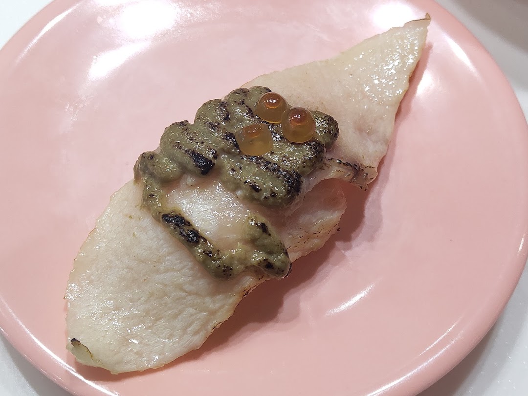 A close-up of a piece of sushi with a green topping and orange roe on a pink plate.