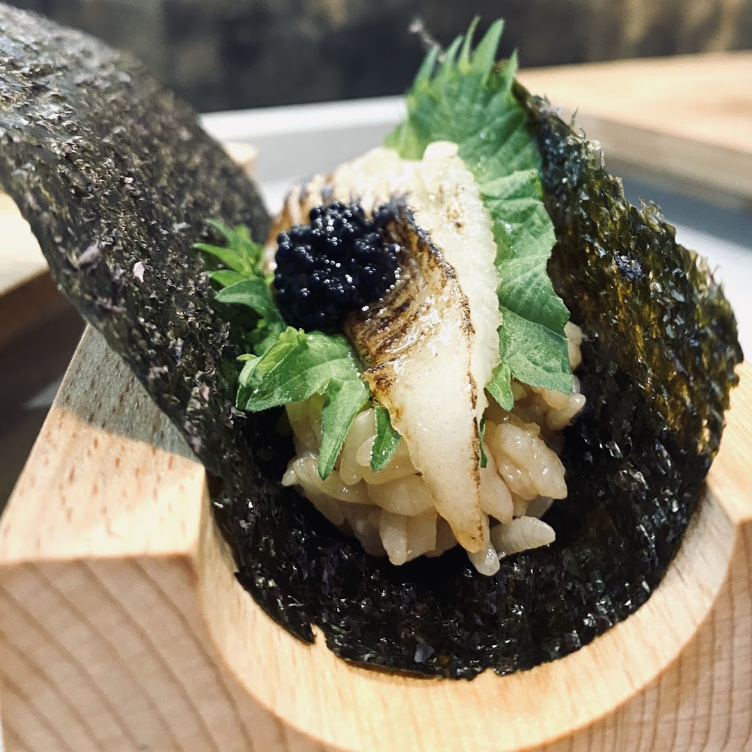 A close-up of a gourmet sushi hand roll featuring seared fish, black roe, and fresh greens, served in a wooden holder.