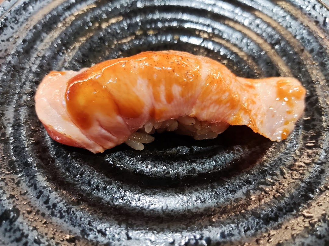 A close-up of a delicious piece of seared salmon sushi with sauce on a dark textured plate.