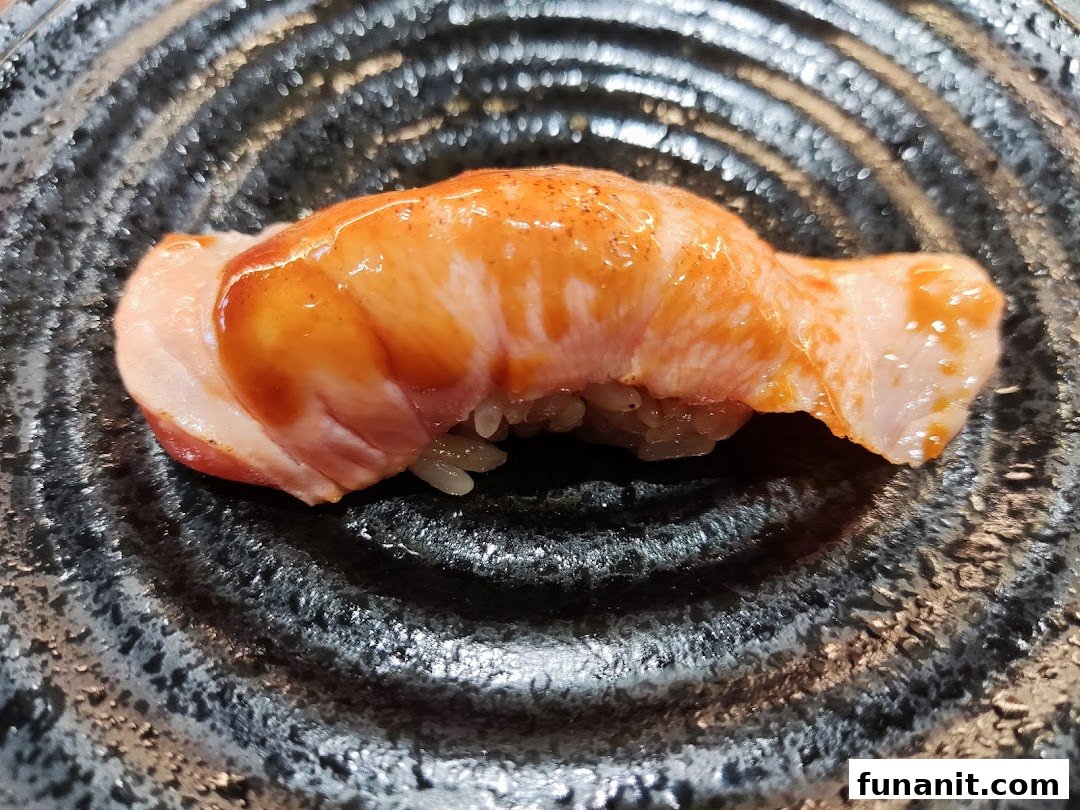 A close-up of a delicious piece of seared salmon sushi with sauce on a dark textured plate.