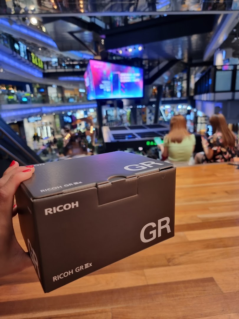 A hand holds a Ricoh GR IIIx camera box inside a bustling shopping mall, likely Funan.