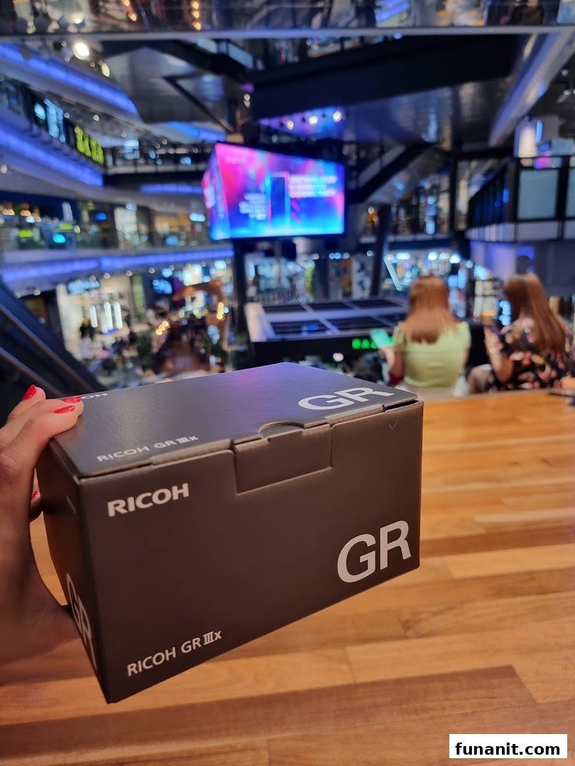 A hand holds a Ricoh GR IIIx camera box inside a bustling shopping mall, likely Funan.