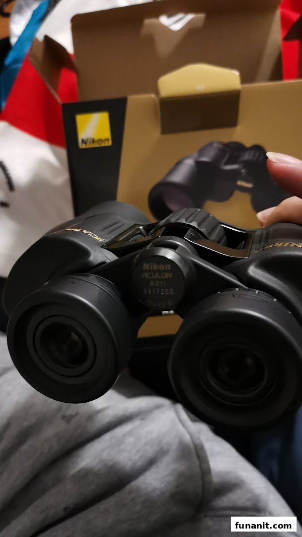 A pair of Nikon Aculon A211 binoculars held, with its product box visible in the background.