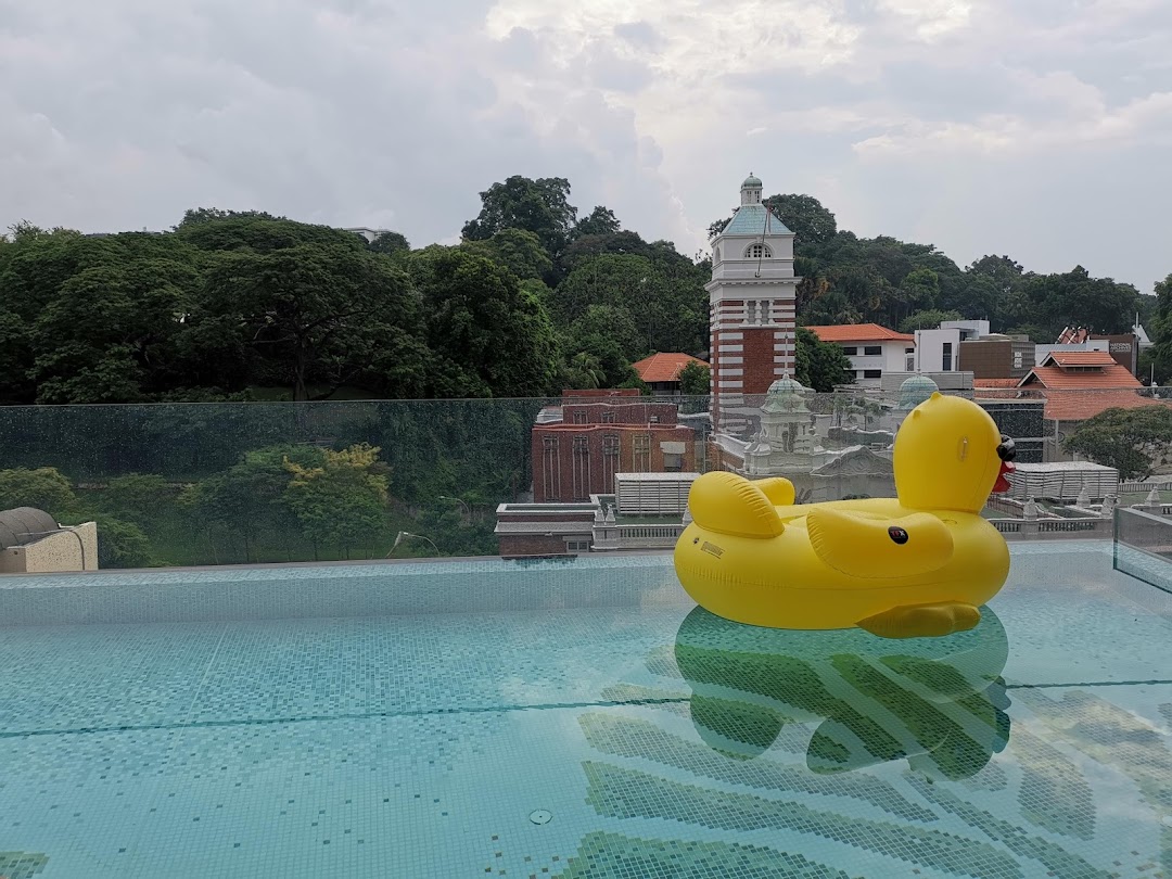 A rooftop swimming pool with a large yellow inflatable duck, overlooking a city skyline and lush greenery.