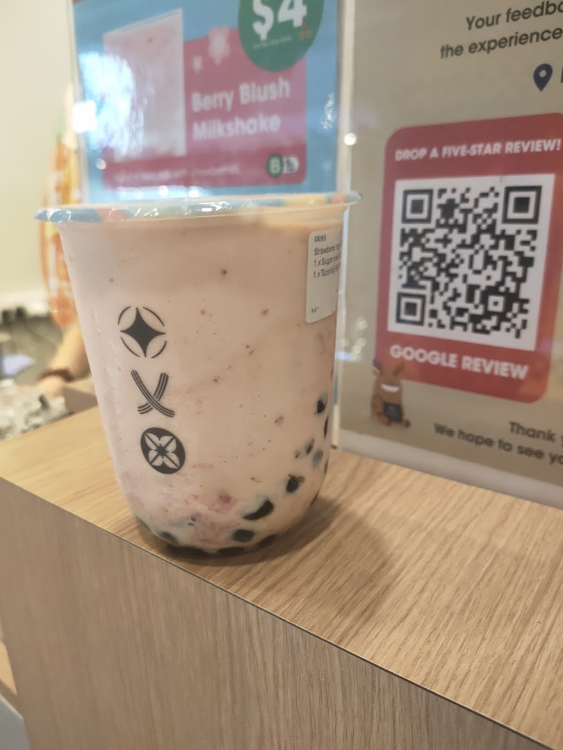 A close-up of a Berry Blush Milkshake with boba pearls on a wooden counter, with promotional signs in the background.