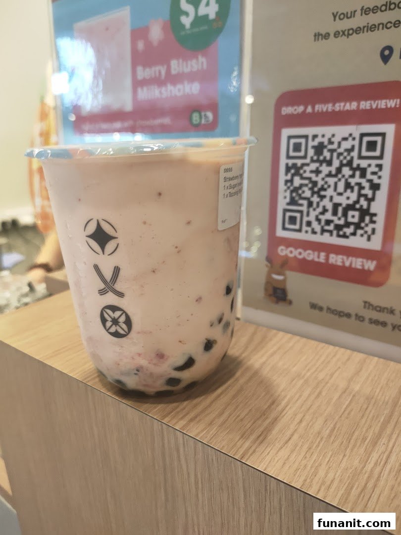 A close-up of a Berry Blush Milkshake with boba pearls on a wooden counter, with promotional signs in the background.