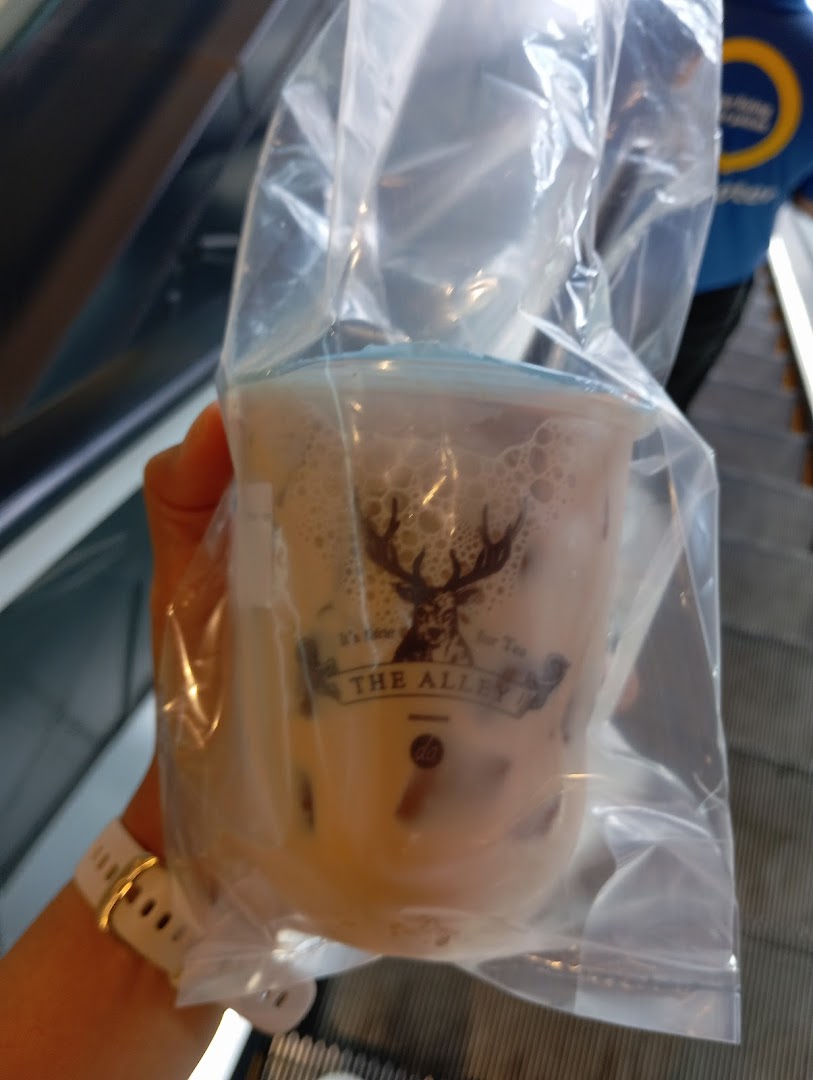 A hand holds a bubble tea drink from The Alley in a plastic bag while on an escalator.