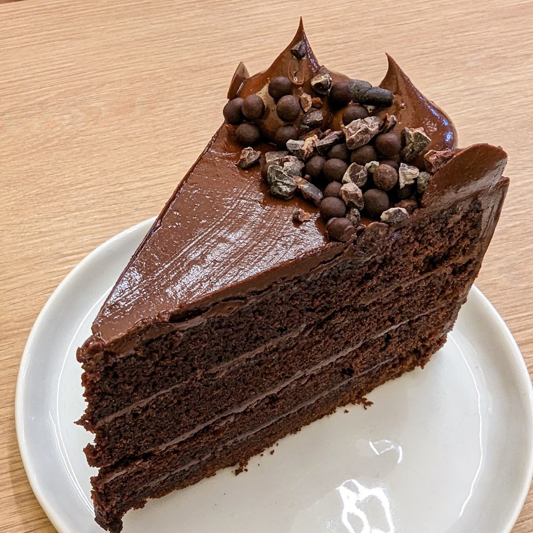 A rich, multi-layered chocolate cake slice topped with ganache, chocolate pearls, and cacao nibs.