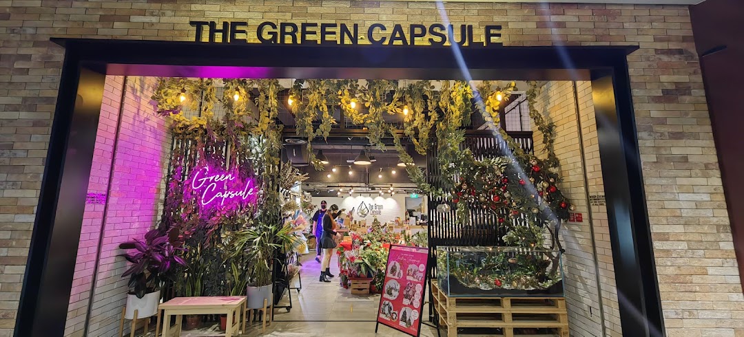 The entrance of 'The Green Capsule' shop, adorned with lush greenery, warm lights, and a vibrant neon sign.