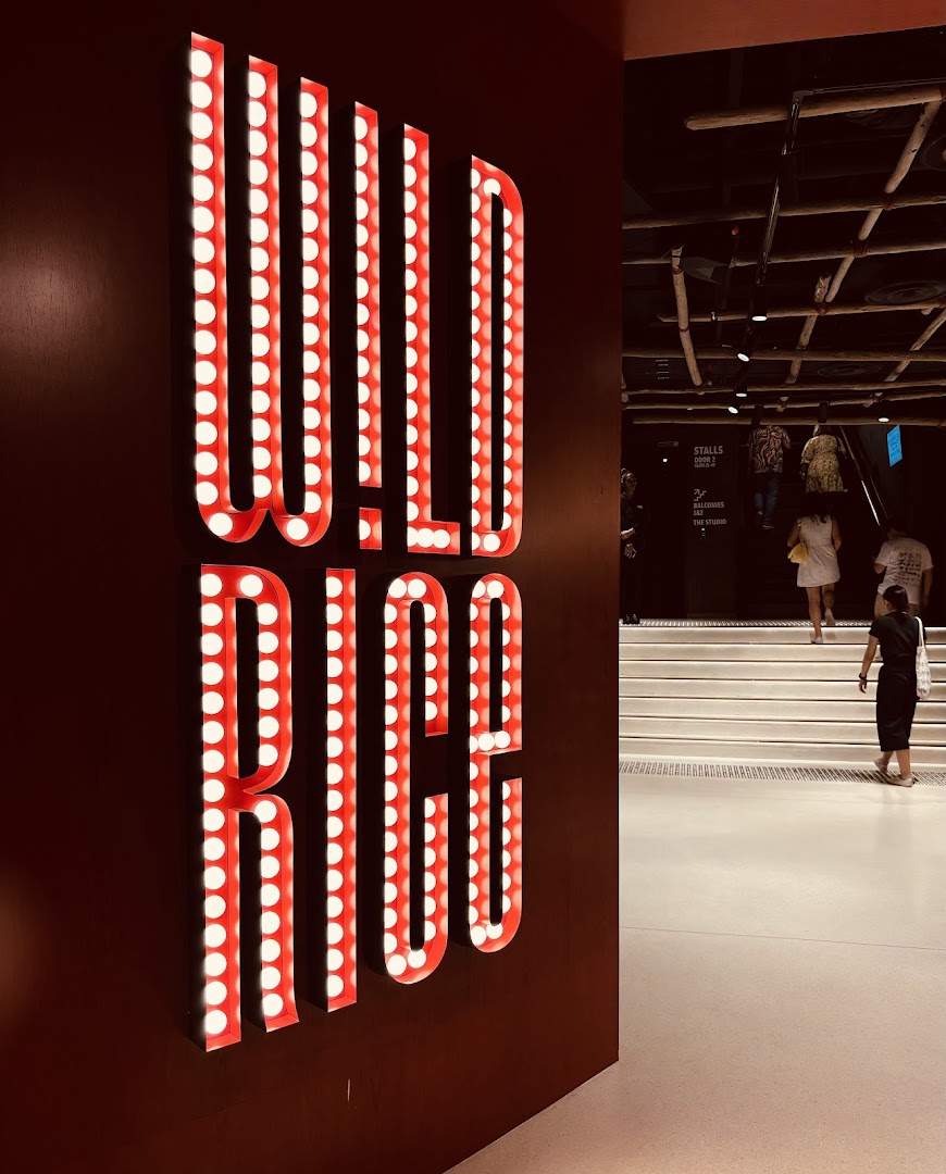 Brightly lit 'WILD RICE' sign at a theatre entrance with people ascending stairs.