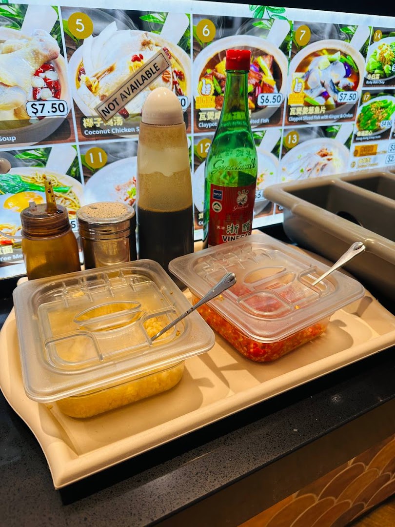 A digital menu board displaying claypot dishes and prices, with condiments on a counter.