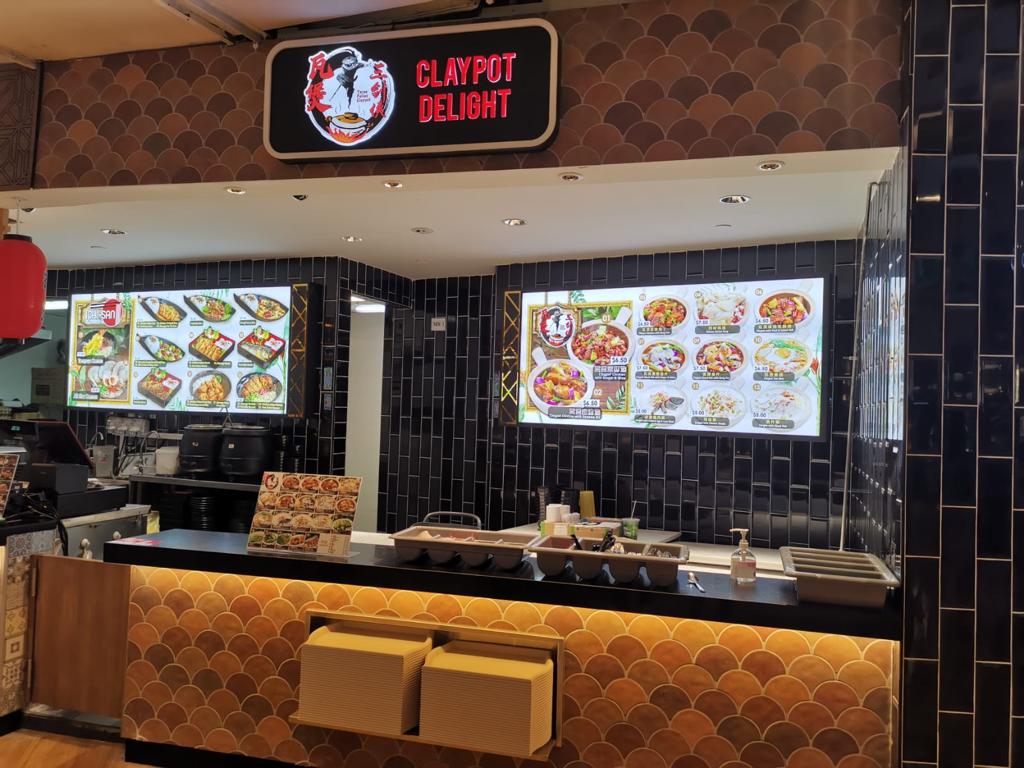 A food stall counter featuring digital menu screens displaying various claypot dishes and pricing.