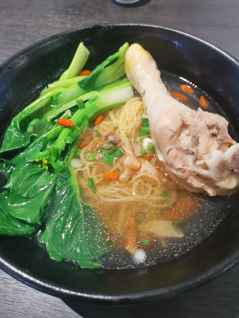A close-up of a delicious bowl of noodles with a chicken drumstick and green vegetables.