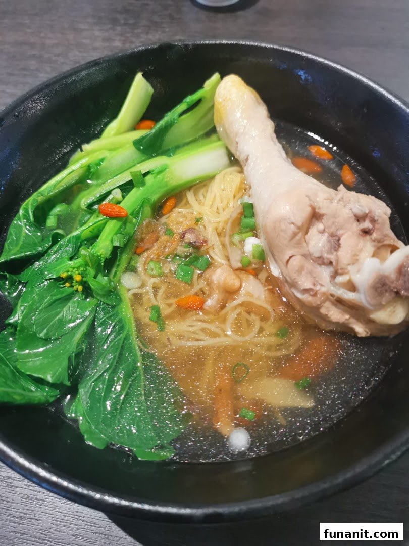 A close-up of a delicious bowl of noodles with a chicken drumstick and green vegetables.