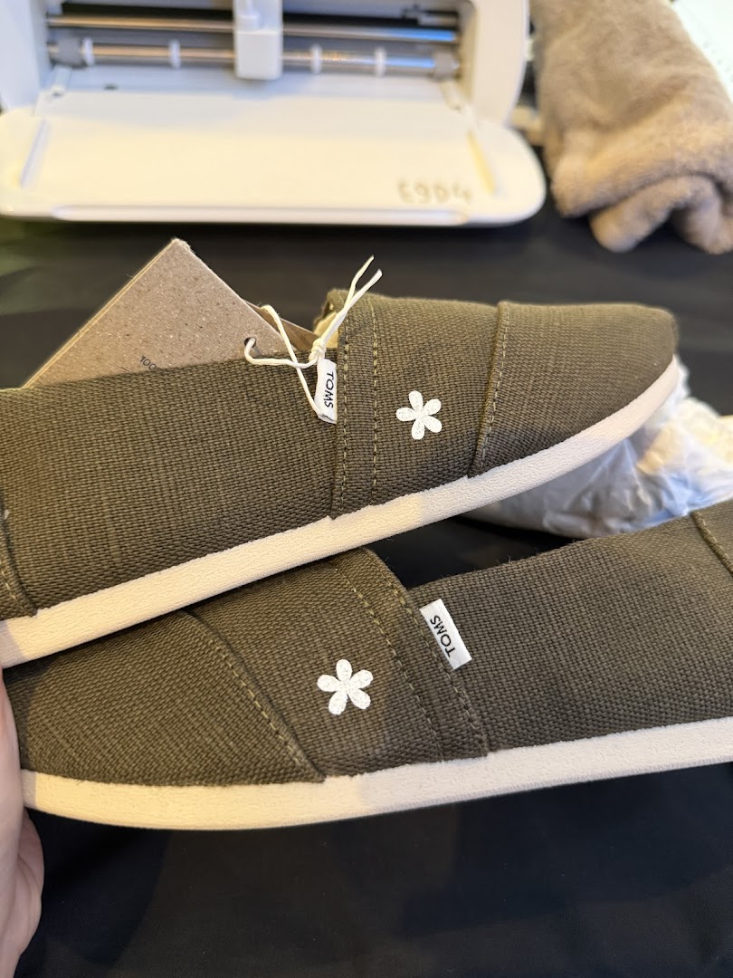 A pair of olive green TOMS shoes with white flower embroidery, showcasing their design.