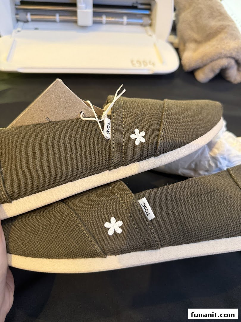 A pair of olive green TOMS shoes with white flower embroidery, showcasing their design.