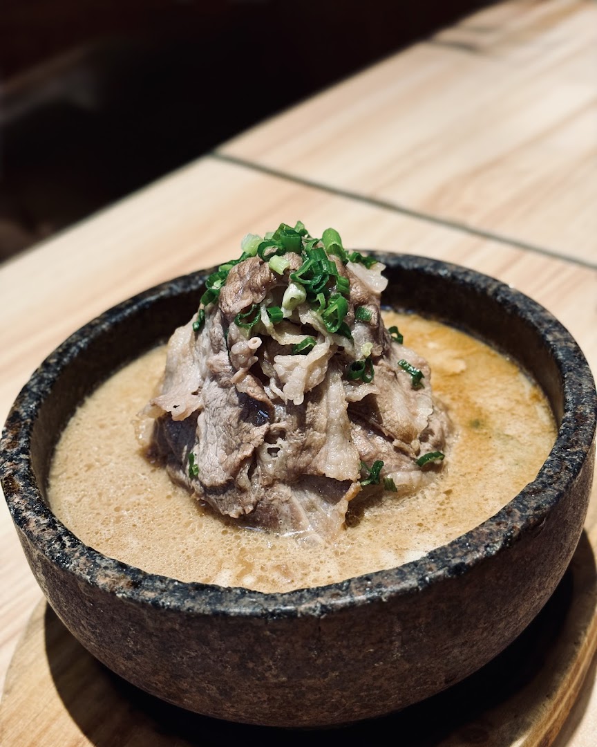 A close-up of a hearty ramen or hotpot dish with thinly sliced beef and green onions in a stone bowl.