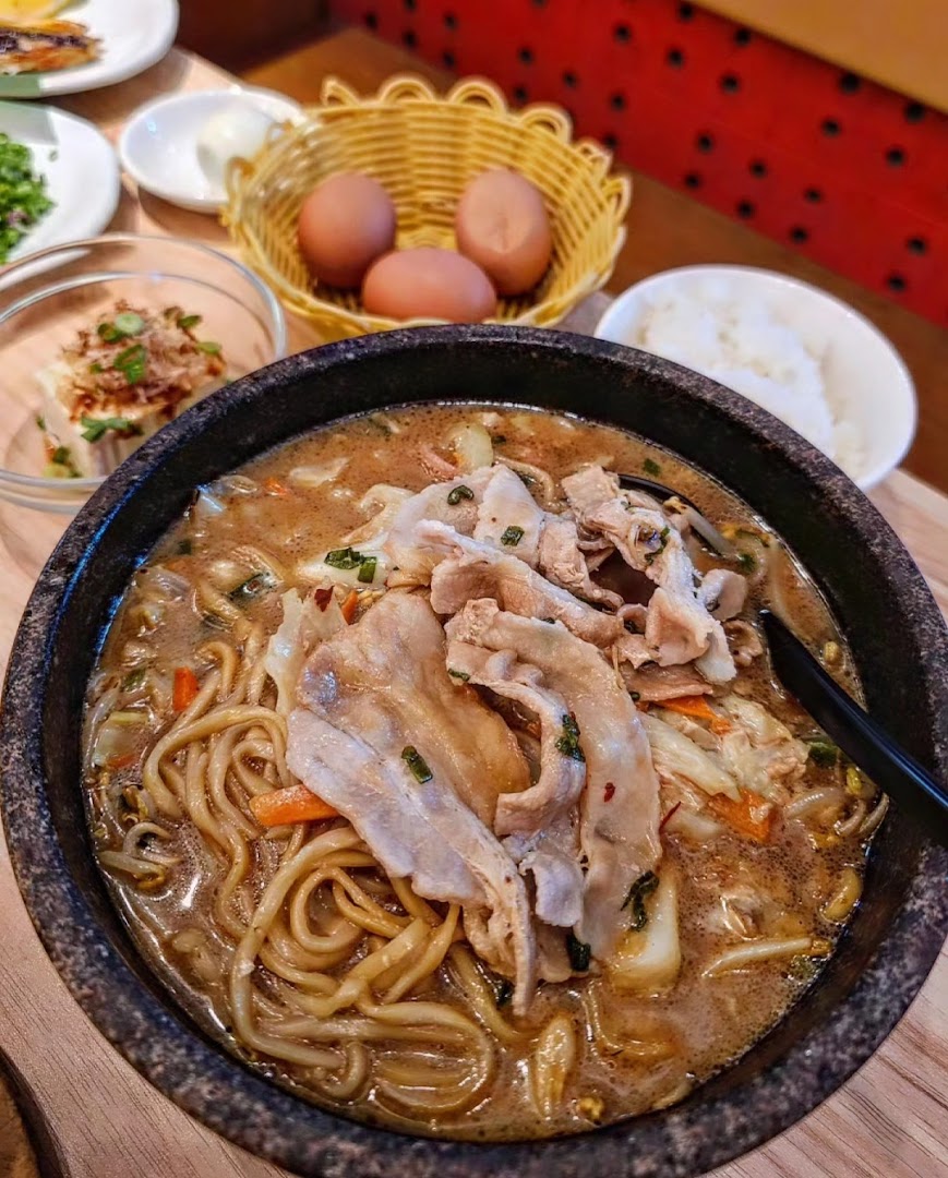 A steaming bowl of Tonkotsu Kazan ramen with pork slices, noodles, and vegetables, served with side dishes.