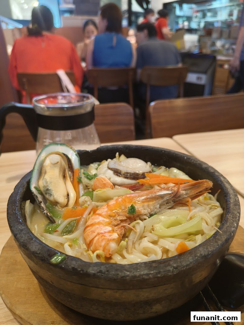 A steaming bowl of seafood ramen with shrimp, mussel, and vegetables on a wooden table in a restaurant.