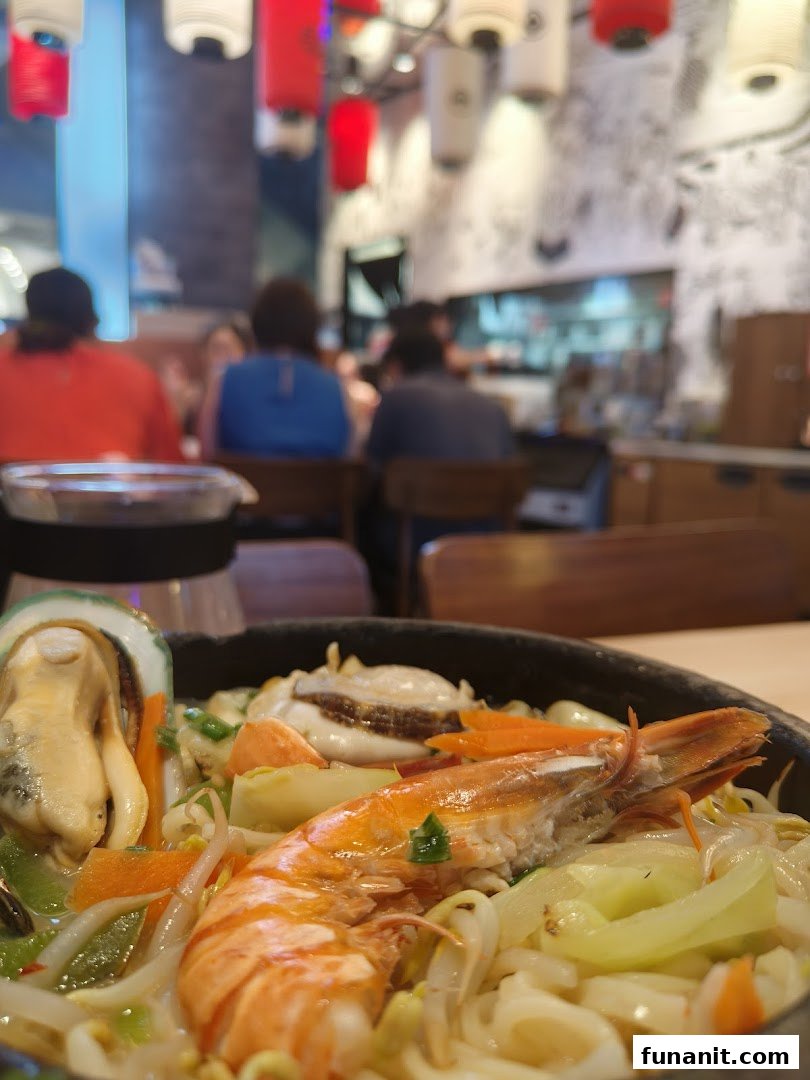 A close-up of a delicious seafood ramen dish with a vibrant restaurant interior and diners in the background.