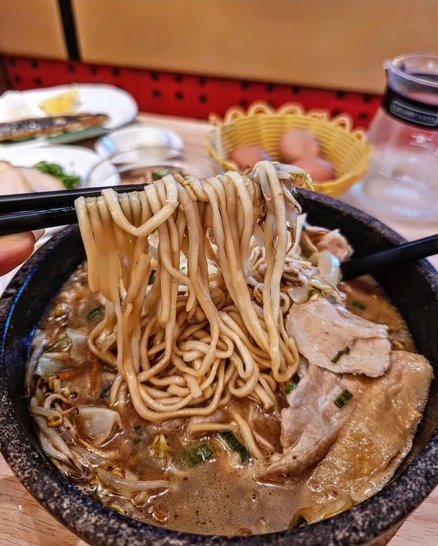 Chopsticks lift thick noodles from a steaming bowl of Tonkotsu Kazan ramen with pork slices and vegetables.