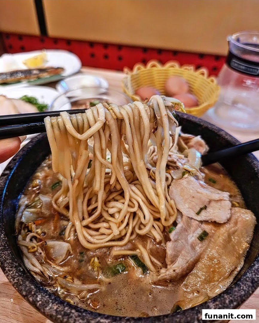 Chopsticks lift thick noodles from a steaming bowl of Tonkotsu Kazan ramen with pork slices and vegetables.