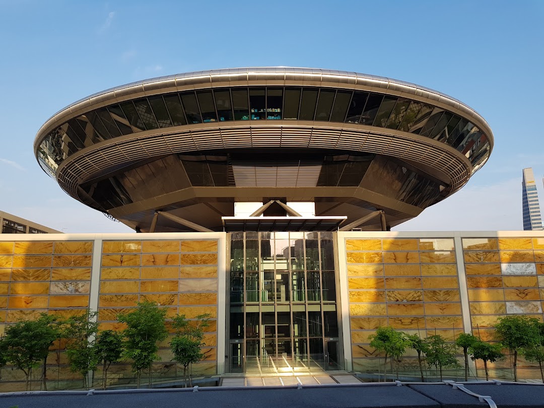 A modern, circular building with glass facades and a golden-patterned wall under a clear sky.