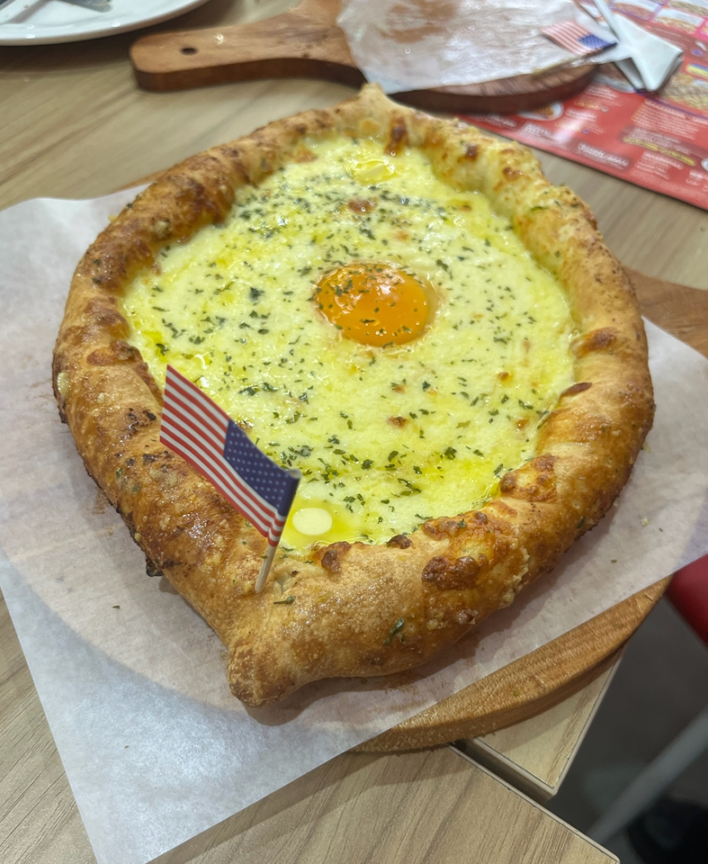 A cheesy, egg-topped pizza-like dish with an American flag pick, served on a wooden board.