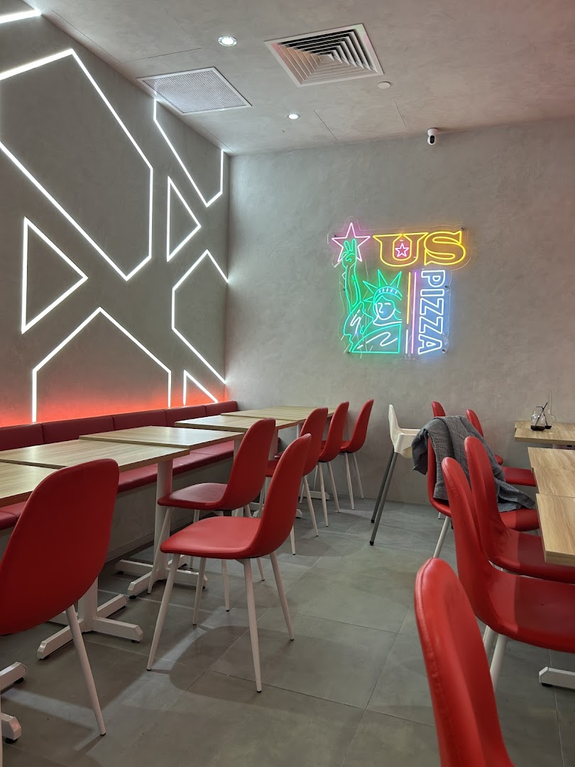 Interior view of US Pizza with red seating, wooden tables, and a vibrant neon logo.
