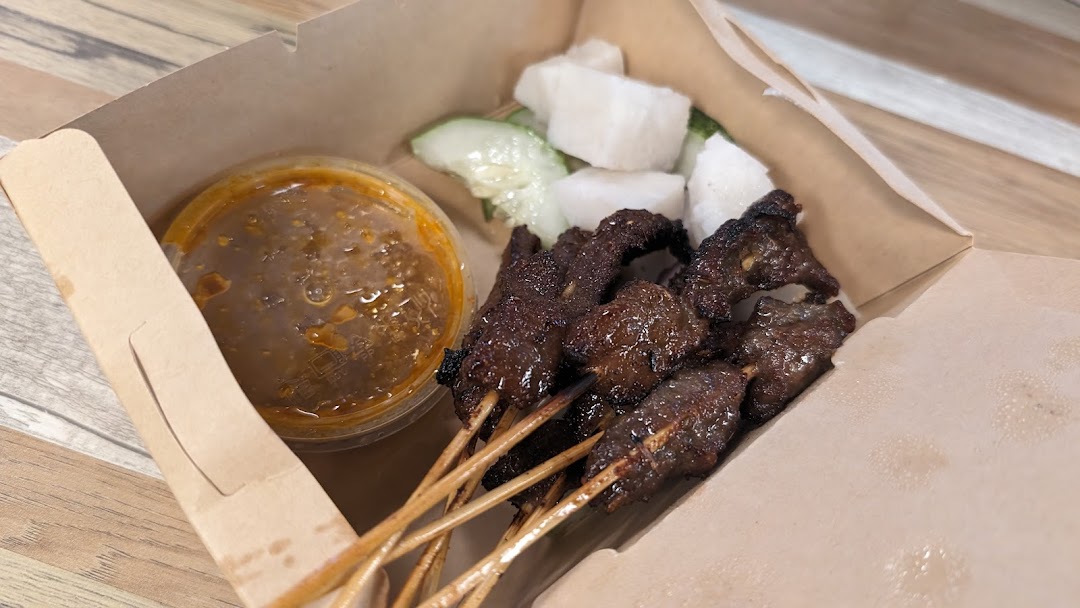 A takeaway box containing grilled satay skewers, peanut sauce, rice cakes, and cucumber slices.
