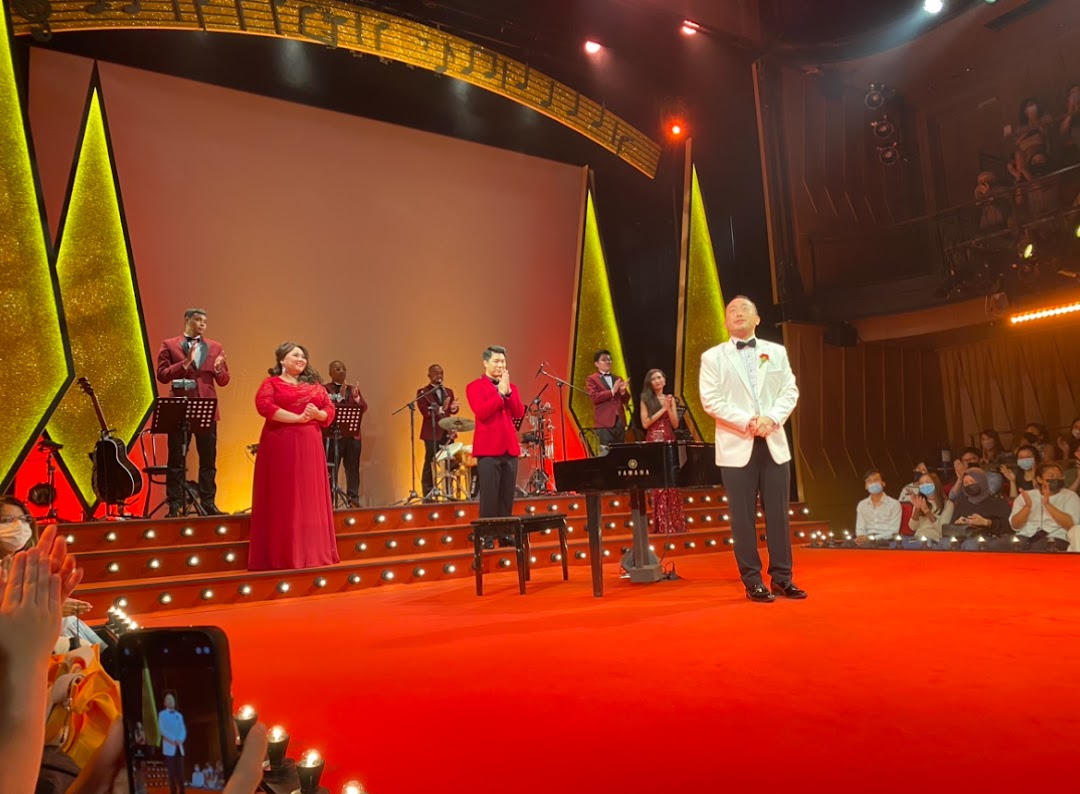 A vibrant stage performance featuring singers, musicians, and a grand piano, with an audience watching.