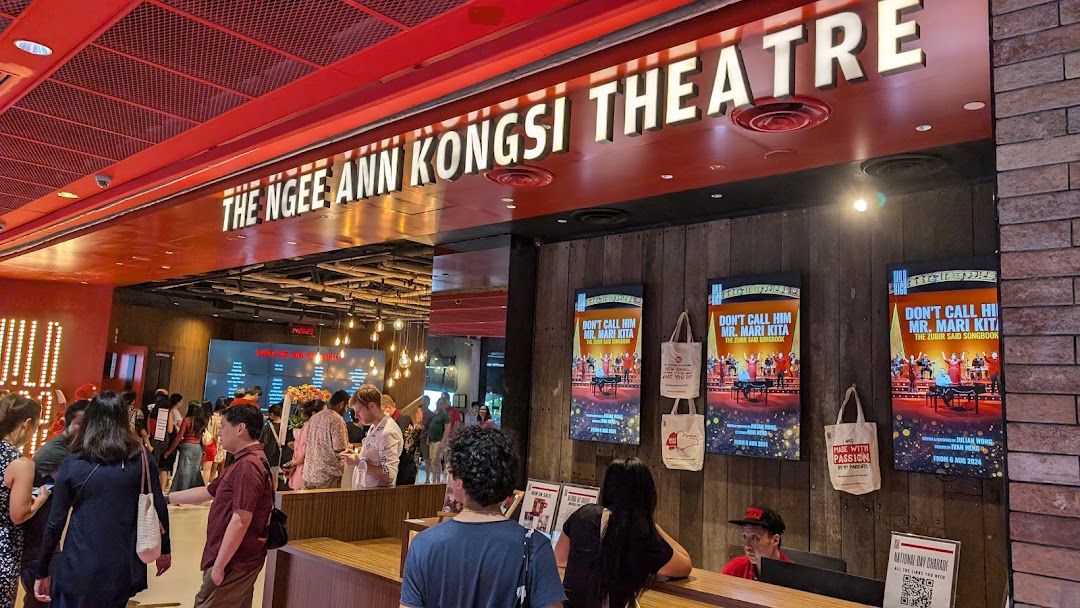 A bustling theatre lobby with patrons, a reception desk, and promotional posters for upcoming shows.
