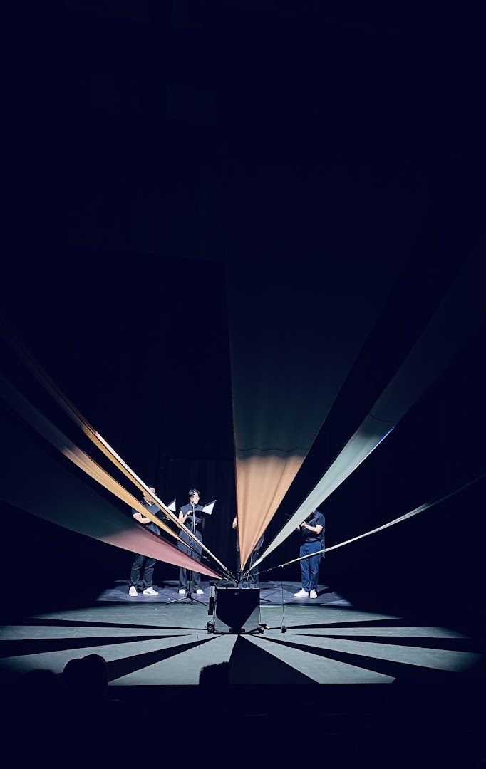 Performers on a dark stage with colorful fabric strips creating a radial pattern and dramatic lighting.