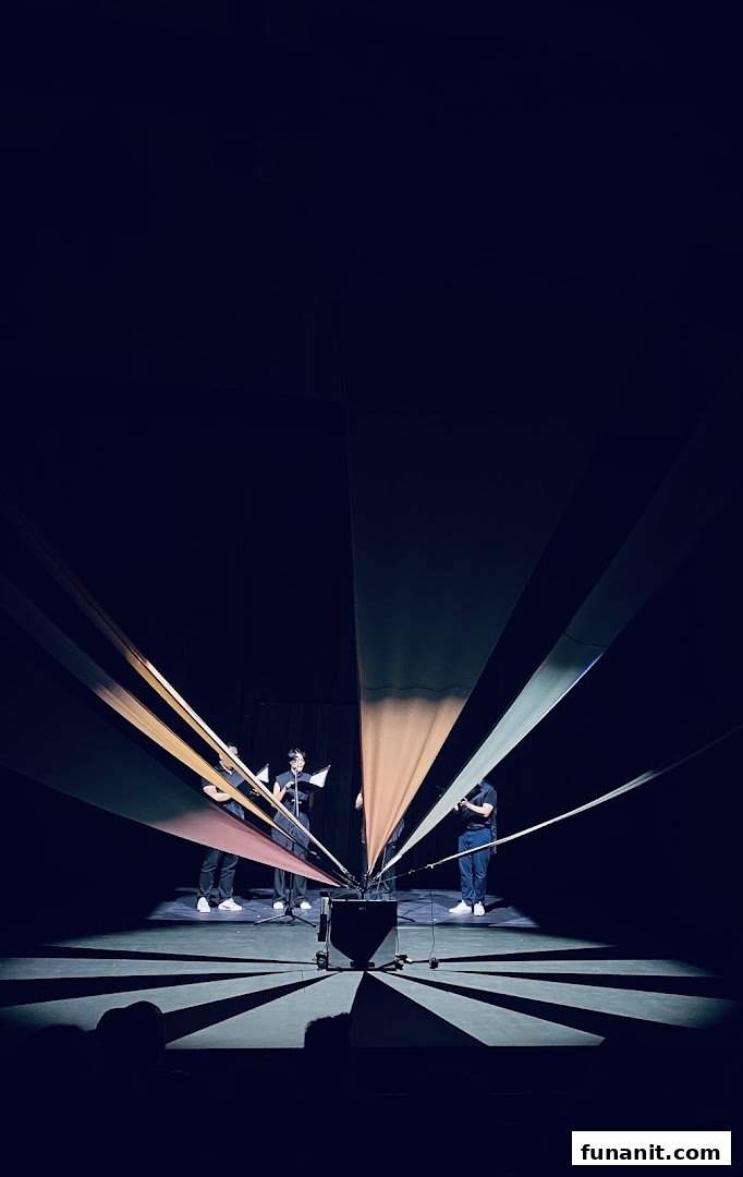 Performers on a dark stage with colorful fabric strips creating a radial pattern and dramatic lighting.